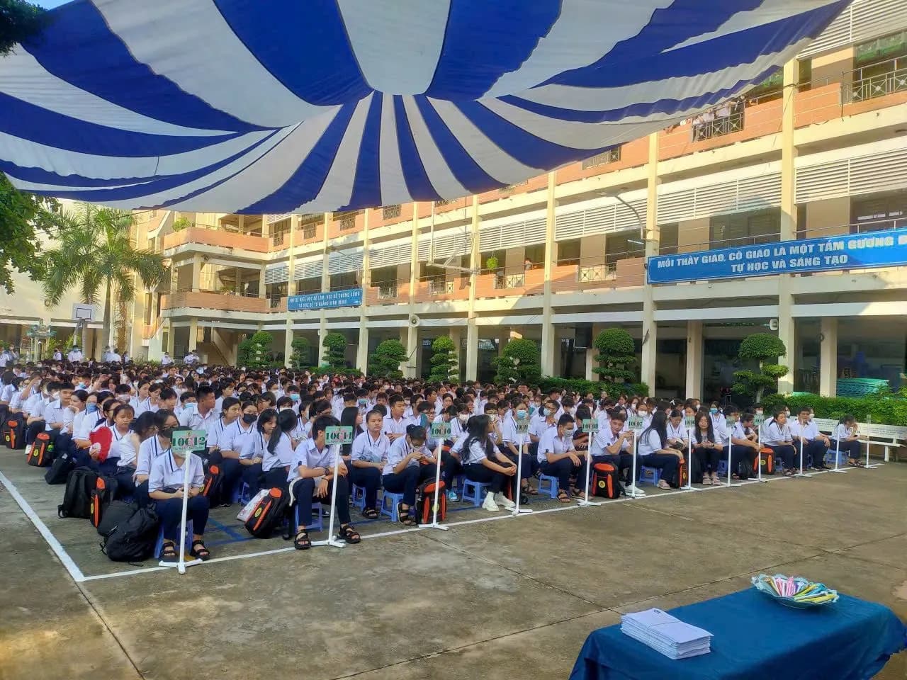 Học sinh ngồi kín sân trường dưới tấm bạt che nắng, mỗi lớp một bảng tên