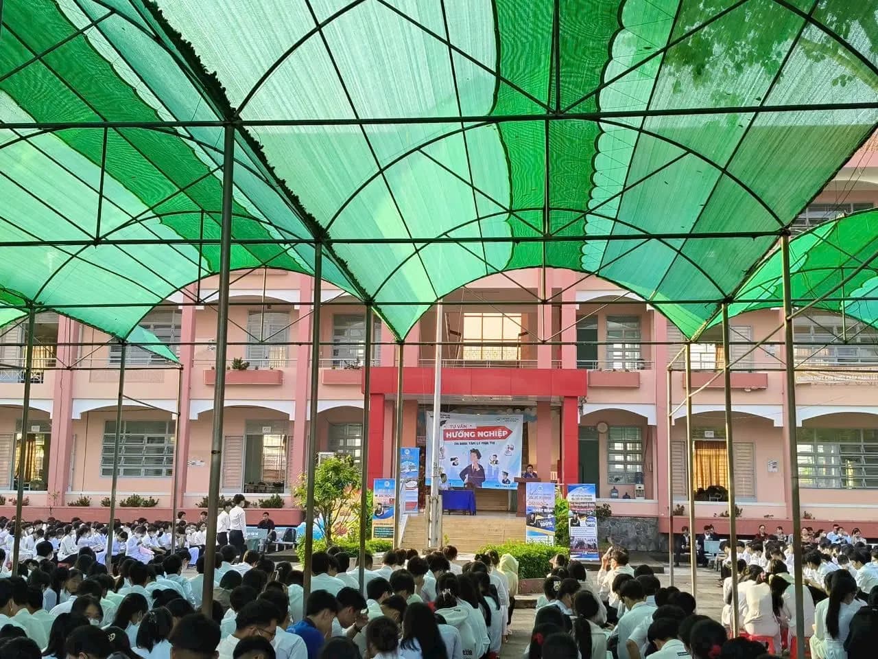 Học sinh ngồi kín sân trường dưới vòm lưới che nắng, hướng về sân khấu